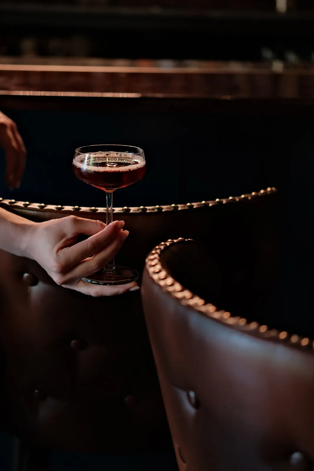 Guest holding a classic cocktail in a coupe glass between leather bar chairs at Sippers
